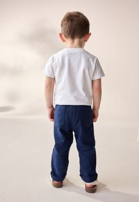 Hvidt bomulds-T-shirt og mørkeblå bukser af denim med opbuk, baglommer, båret af et barn, der står set bagfra.