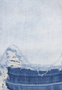 Ljusblå jeansfabric med fransade kanter, med en sliten yta med råa sömmar och en texturerad finish. Närbild av materialets väv.