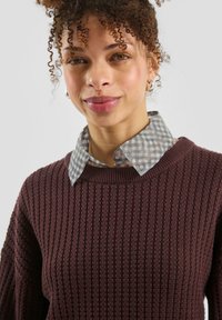 Bordowy sweter z teksturowanym splotem o okrągłym dekolcie, noszony na jasnoszarej wełnianej koszuli w kratę z kołnierzykiem. Z mankietami wykończonymi prążkowanym ściągaczem.