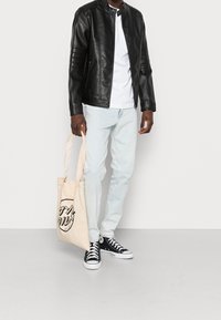 Veste en cuir noire avec fermeture pression, t-shirt blanc, jean bleu clair et baskets noires. Équipé d'un sac fourre-tout beige avec imprimé graphique.