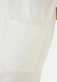 Helles Stoffkleidungsstück mit einer großen seitlichen Tasche, die zarte florale Stickerei aufweist. Glatte Textur und dezentes Rippmuster über das gesamte Kleidungsstück.