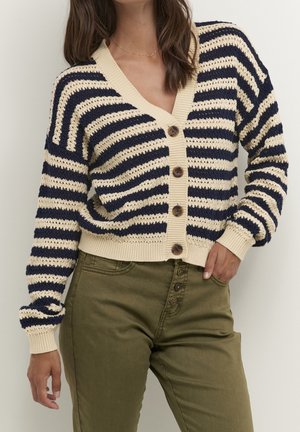 Kvinde iført en creme- og marineblåstribet cardigan med knapper og olivengrønne højtaljede bukser, med hånd i lommen.