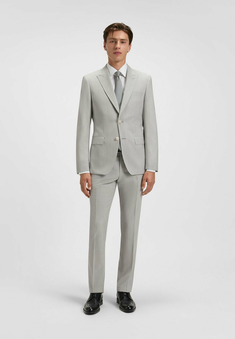 Jasnoszary garnitur dopasowany, z jednorzędową marynarką, klapami w kształcie notcha, dwiema przednimi kieszeniami oraz spodniami prostymi. W zestawie z białą koszulą i krawatem.