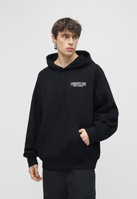 Nuori mies seisoo yllään musta Carhartt WIP -huppari, jossa on etutasku, katsoo sivulle vaaleanharmaan yksivärisen taustan edessä.