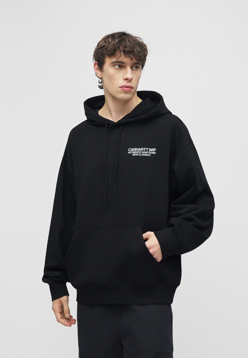 Giovane in piedi che indossa una felpa nera Carhartt WIP con tasca frontale, guarda di lato contro uno sfondo grigio chiaro uniforme.