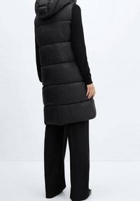 Veste matelassée noire sans manches avec capuche, présentant un matelassage horizontal, une coupe mi-longue et une texture lisse et brillante. Portée par-dessus un haut à manches longues noir.