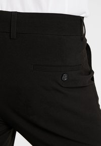 Schwarze, maßgeschneiderte Hose mit glatter Stofftextur, flacher Vorderseite und einer einzelnen, mit Knopf verschlossenen Gesäßtasche. Schlanke Passform mit Gürtelschlaufen.