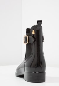 Botte noire brillante à la cheville avec panneaux élastiques, accent de boucle dorée et un talon texturé. Matériau lisse avec une silhouette élégante.