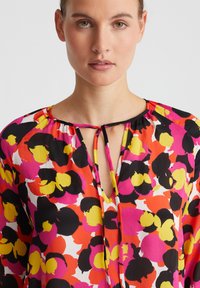 Helle Blumenbluse mit einem Mix aus Rosa, Gelb und Schwarz. Runder Ausschnitt mit einer Bindeschleife. Leichtes, weiches Material.