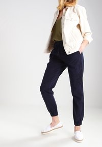 Veste légère beige sur un haut olive ; pantalon à taille haute bleu marine avec des poignets élastiques. Le modèle porte des espadrilles blanches.
