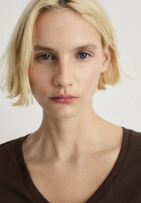 Portrét osoby s krátkymi, rovnými blond vlasmi a modrými očami, ktorá má na sebe hnedý vrchný diel s výstrihom do V, s hladkou textúrou a jednoduchým dizajnom.