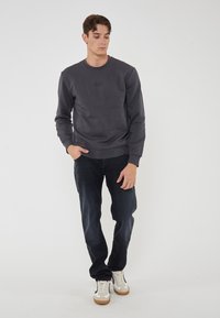 Felpa girocollo grigia scura con un logo discreto, abbinata a jeans scuri e sneakers bianche. L'outfit ha un aspetto casual e lineare.