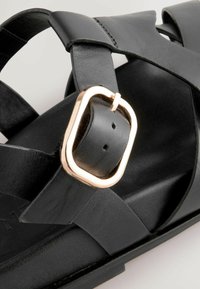 Sandale en cuir noir avec des lanières croisées, texture lisse et une boucle en métal doré pour un ajustement réglable. Design de semelle plate.