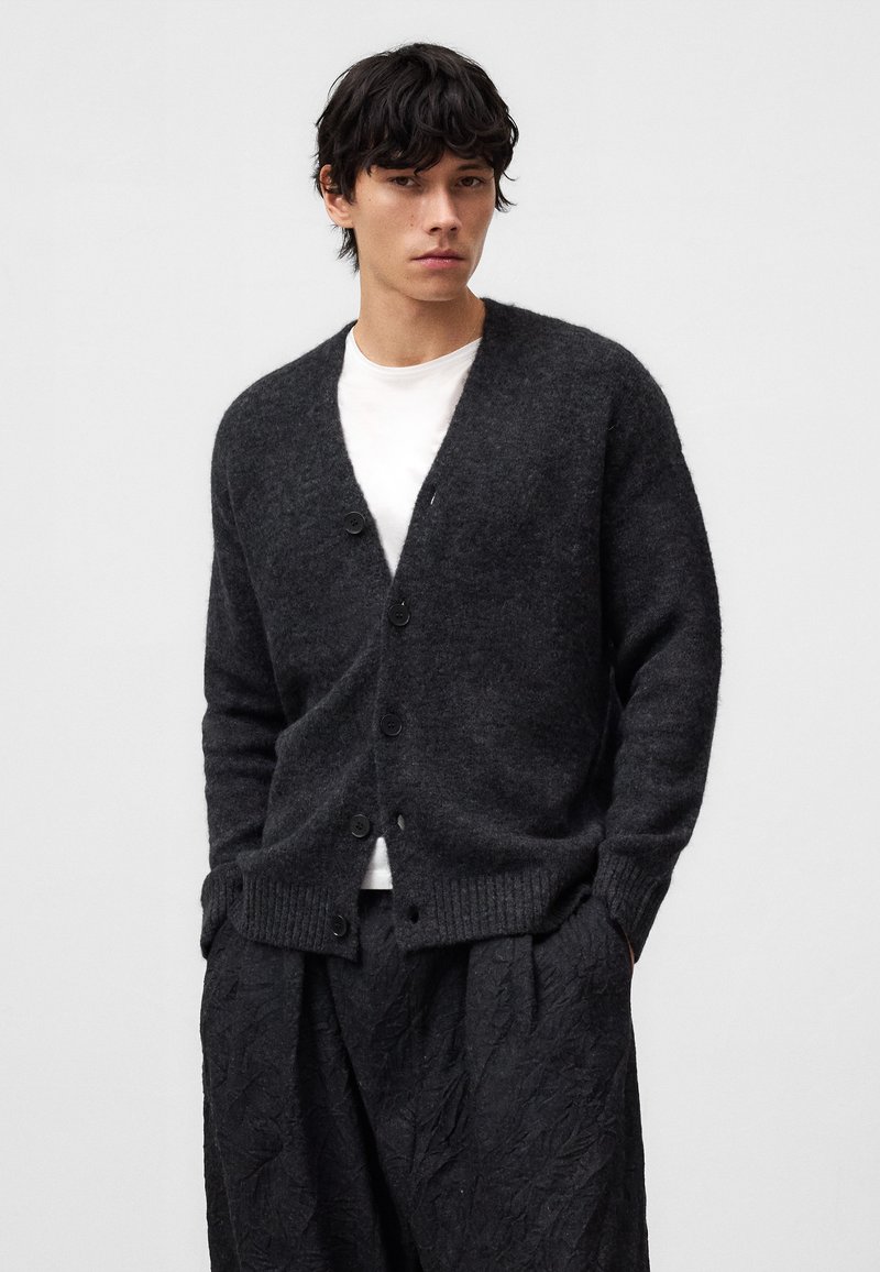 Mörkgrå stickad cardigan med knappframsida, ribbade manschetter och nederkant, i kombination med lösa svarta texturerade byxor. Enkelt design, mjuk textur.