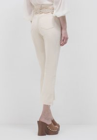 Una donna che indossa pantaloni tagliati crema con orli sfilacciati, una camicia bianca, una cintura decorativa e sandali aperti con tacco a piattaforma marroni, rivolta di lato.