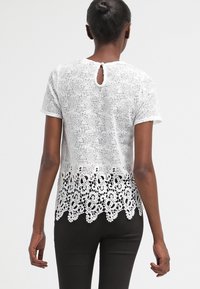 Top bianco in pizzo con una parte superiore testurizzata e un orlo decorativo a scallop con motivi floreali. Maniche corte e chiusura a forma di chiave sul retro.
