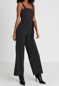 Svart prickig byxdress med fyrkantig halsringning, bred bendesign och midja med knytning. Tyget verkar lätt och har en jämn textur.