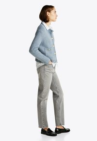 Gilet tricoté texturé bleu clair avec des boutons dorés, deux poches avant, porté sur une chemise blanche, associé à un pantalon droit gris et des mocassins noirs.