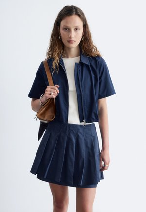 Jeune femme aux cheveux bouclés portant un haut blanc, une veste bleue marine à manches courtes avec fermeture éclair, une jupe plissée bleu marine et tenant un sac à épaule marron.