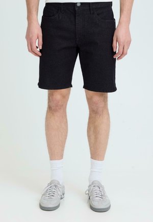 Mann iført svarte jeansshorts, hvite ankelsokker og lyse grå joggesko, stående mot en ensfarget hvit bakgrunn.