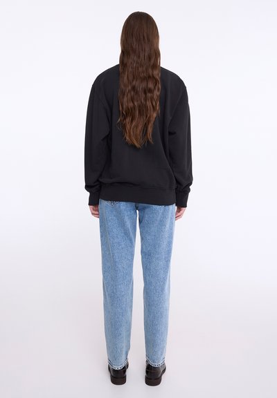 Fekete kapucnis pulóver hosszú ujjal, laza szabásban, bordás mandzsettával, világoskék, laza szabású farmernadrággal és fekete cipővel párosítva.