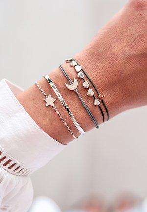 Sæt af fem armbånd på et håndled: sølvarmbånd, mørke snorarmbånd med hjerteformer, en halvmåne og en stjernecharmer.