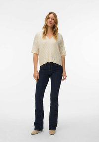 Lichtbeige gebreide trui met V-hals en korte mouwen, met een ruitpatroon, gecombineerd met donkerblauwe wijde jeans en beige schoenen.