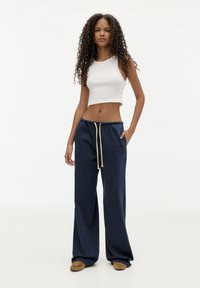 Ung kvinde med langt krøllet hår iført en hvid ærmeløs crop top, marineblå vada bukser med snøre og brune sko, stående.