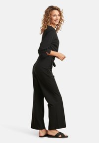 Schwarzer Jumpsuit mit Dreiviertelärmeln, Taillenknoten und weitem Bein. Getragen mit schwarzen, geflochtenen Schlittensandalen vor einem hellen Hintergrund.