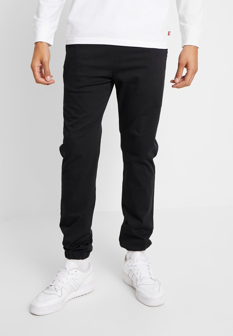 Homme portant un pantalon de jogging noir avec des poignets élastiques et des baskets blanches, debout devant un fond clair uni.