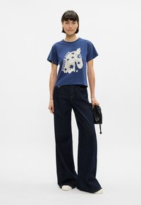 Ναυτικό μπλε κοντό μπλουζάκι με άσπρο γραφικό σχέδιο, σε συνδυασμό με χαλαρούς σκούρους μπλε τζιν. Το μοντέλο κρατάει μια μικρή μαύρη τσάντα.