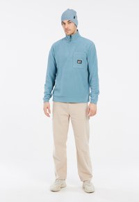 Šviesiai mėlynas fliso puloveris su užtrauktuku kaklo srityje ir krūtinės kišene, derinamas su smėlio spalvos kelnėmis ir šviesiai pilkais sportbačiais. Modelis vilki atitinkamus kepuraitę.