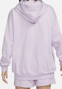 Osoba oblečená v lehce fialové oversized mikině a ladících šortkách, zobrazená zezadu s uvolněnýma rukama po stranách.