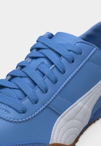 Baskets bleues avec des lacets bleus texturés, une couture détaillée et une bande latérale blanche, présentées en gros plan sur un fond clair.