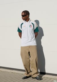 Hvid poloshirt med grønne detaljer og et emblem med Italiens flag, beige posede bukser og sorte sporty sko, accessoriseret med solbriller.