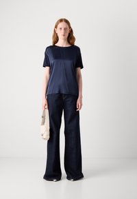 Top corto a maniche corte in satin blu navy abbinato a jeans in denim a gamba larga di colore blu scuro. Il modello tiene una borsa color crema con un design strutturato.