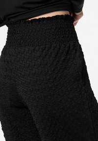 Melnas teksturētas bikses ar augstu, saspiestu vidukļa daļu. Audumam ir izcelta raksta tekstūra, kas piešķir unikālu taustes kvalitāti un izskatu.