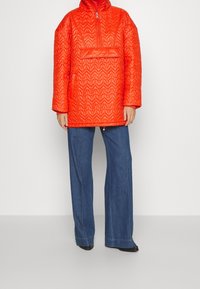 Pikowana pomarańczowa bluza z wysokim kołnierzem, przednią kieszenią i ściągaczem u dołu, w zestawieniu z szerokimi, niebieskimi dżinsowymi spodniami.