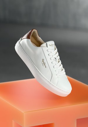 Sneaker bianco in pelle con tallone in pelle marrone texturizzato, chiusura con lacci, forma liscia e dettaglio del logo sul lato, posizionato su un piedistallo arancione.