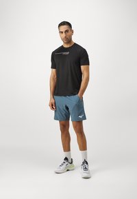 Zwart sportshirt met korte mouwen en logo, gecombineerd met blauwe shorts met zijzakken, en witte sneakers met marineblauwe en groene accentkleuren.