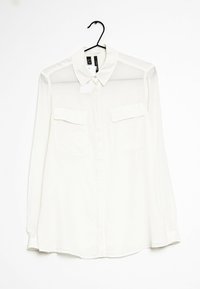 Valkoinen pitkähiha blouse, jossa on kaulus, kaksi rintataskua ja nappi-etuosa. Läpinäkyvä kangas, jossa on sileä tekstuuri. Ripustettuna mustalta koukulta.