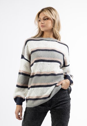 Donna che indossa un maglione a righe largo di colore bianco, grigio, rosa e blu navy, abbinato a jeans grigio scuro, in piedi con una mano in tasca.