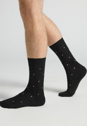 Schwarze Socken mit glatter Textur, die ein Muster aus kleinen roten Punkten aufweisen, die gleichmäßig über den Stoff verteilt sind. Klassisches Crew-Längen-Design.