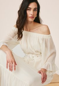 Femme aux longs cheveux foncés portant une robe blanche épaules dénudées avec des manches bouffantes et une large ceinture, assise et regardant la caméra.