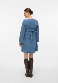 Denim obleka z dolgimi rokavi, zategnjeno pasom in detajlom z vezavo na hrbtni strani. Nosi se s kostanjastimi škornji do kolen, gladke teksture, svetlo modre barve.