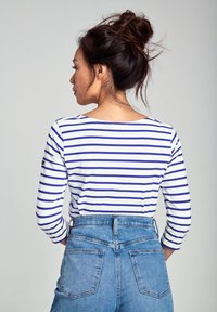 Maglietta bianca a maniche lunghe con righe blu, scollatura rotonda, indossata con jeans di denim a vita alta con tasche posteriori e cuciture visibili.