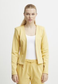 Žlutý blazer s hladkou strukturou, otevřeným předním designem a dlouhými rukávy, spárovaný s odpovídajícími žlutými kalhotami s elastickým pasem.
