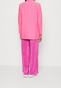 Růžový blazer se straight střihem a klopovým límcem v kombinaci s plisovanými, splývavými růžovými kalhotami. Materiál vypadá hladce s mírně lesklou texturou.