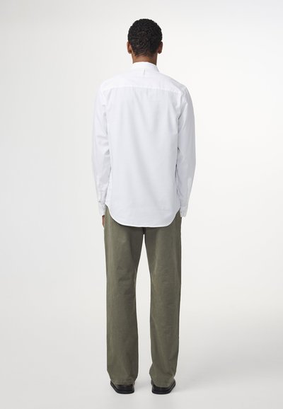 Witte longsleeve shirt met knoopmanchetten, gebogen zoom en een rugpasdetail, gecombineerd met loszittende olijfgroene broek en zwarte schoenen.