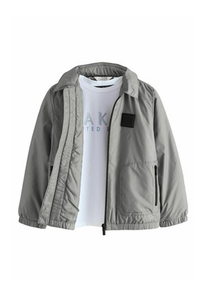 Sivi lagani jakna s patentnim zatvaračem, bočnim džepovima i elastičnim manžetama, složena preko bijele majice s plavim grafičkim printom.